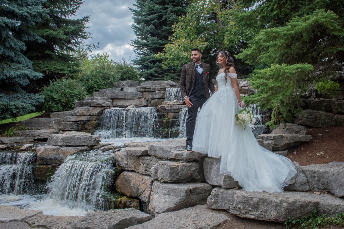 A smiling bride in a white gown and a groom in a suit stand beside a stone waterfall in a forest garden.