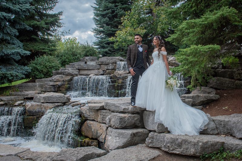 A smiling bride in a white gown and a groom in a suit stand beside a stone waterfall in a forest garden.