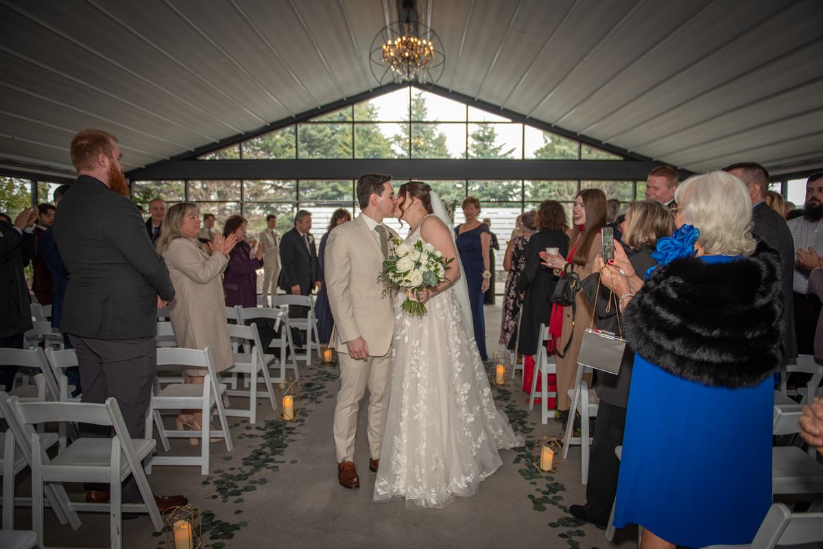 Bride and groom kiss at the end of their aisle, surrounded by guests and illuminated by natural light and candles.