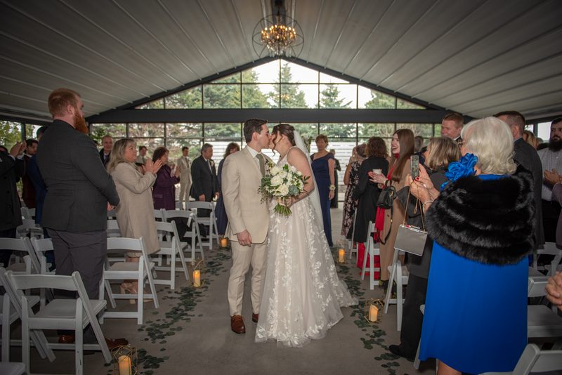 Bride and groom kiss at the end of their aisle, surrounded by guests and illuminated by natural light and candles.