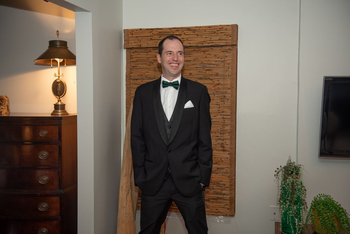 A man in a tuxedo stands by a wooden dresser, with a vintage lamp and bamboo wall panel nearby, in an indoor setting.