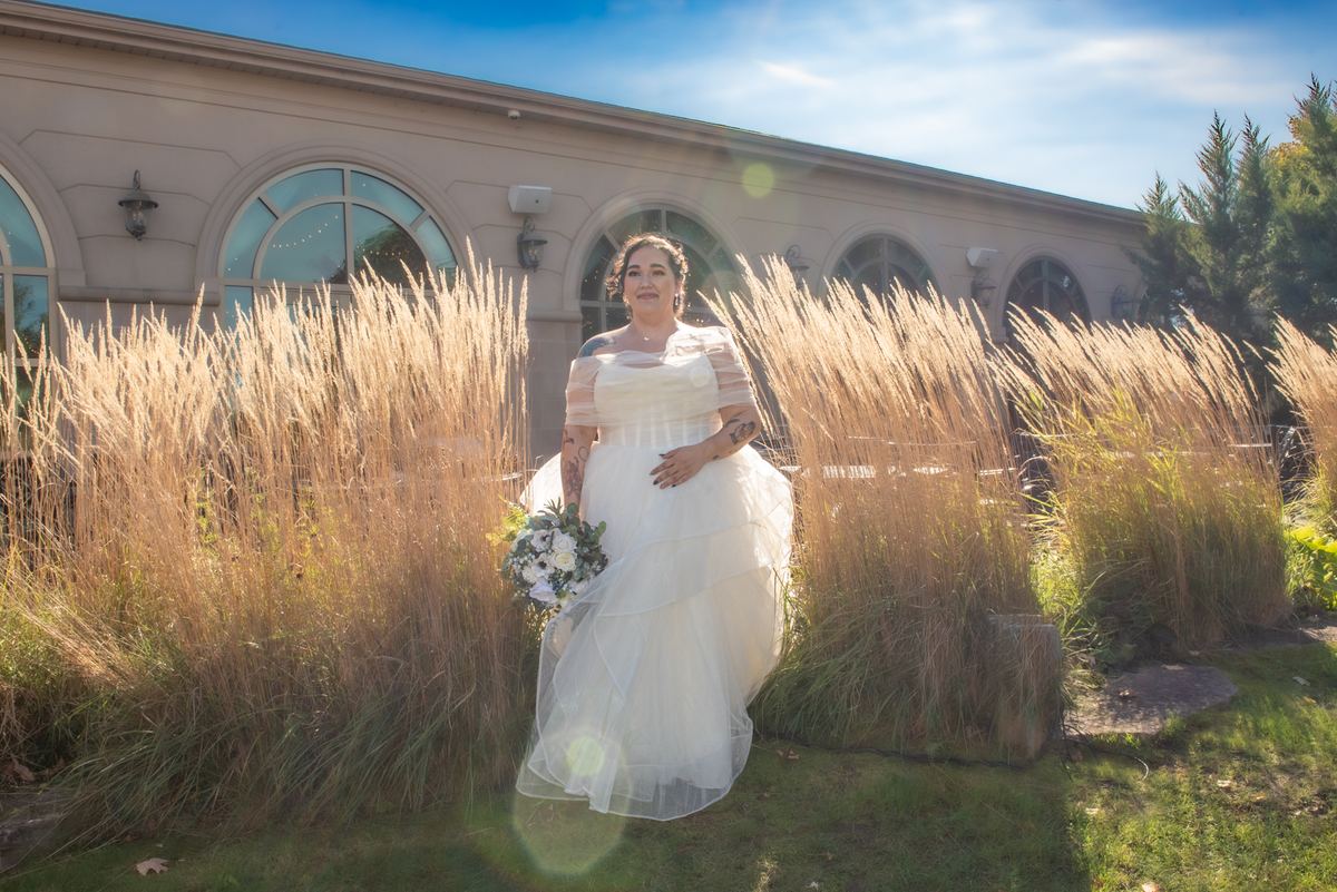 A bride smiles with a bouquet in a sunlit garden, framed by golden grasses and an arched window building.