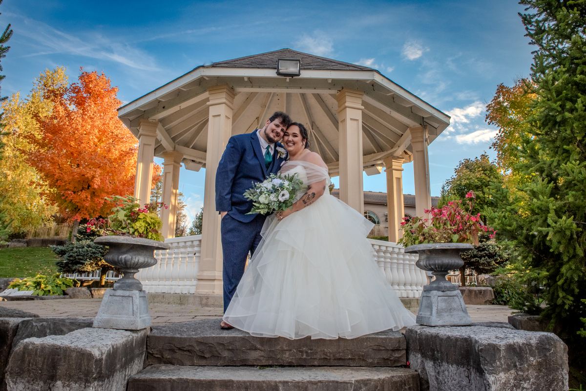 A newlywed couple poses on stone steps beneath a gazebo, surrounded by autumn leaves and smiling at the camera.