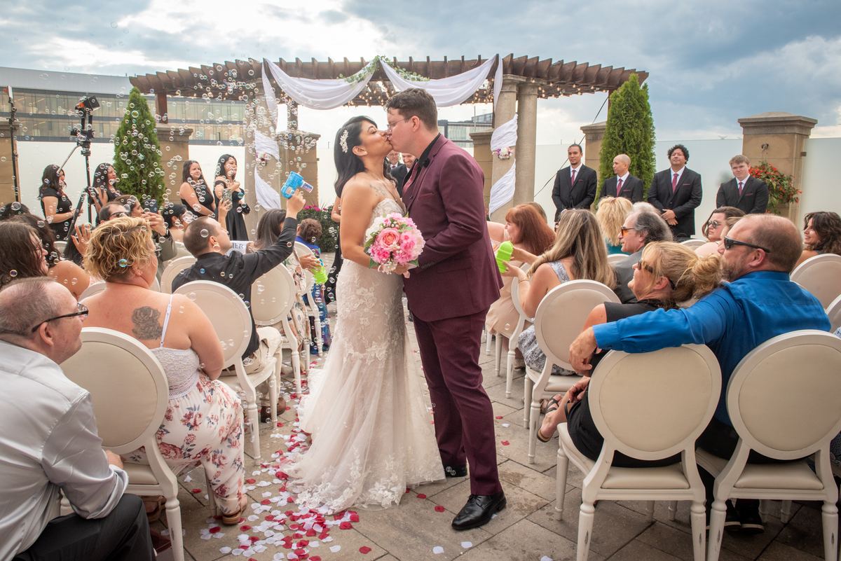 Bride and groom kiss at outdoor ceremony surrounded by guests holding bubble wands and scattering petals.