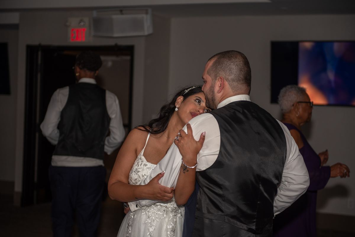 A bride and groom dance closely in a softly lit room, their faces nearly touching as guests mingle nearby.