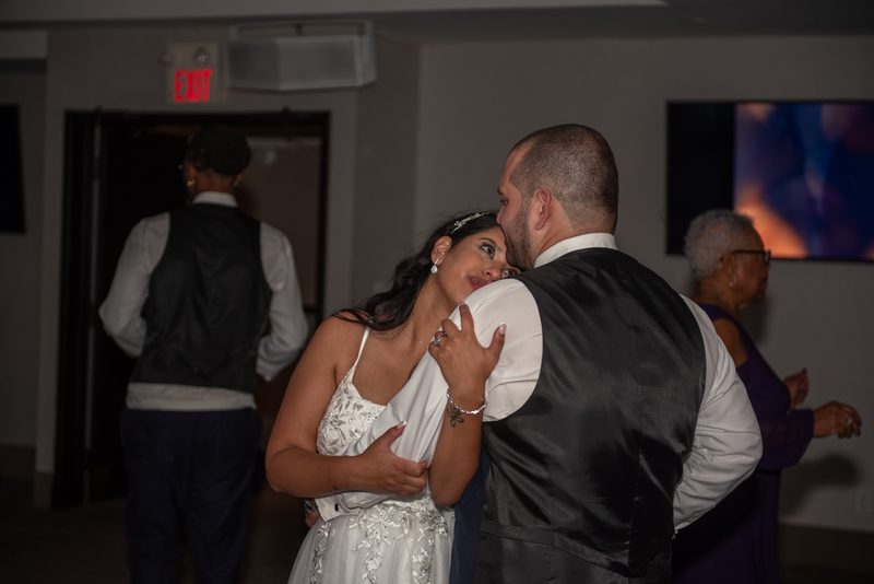 A bride and groom dance closely in a softly lit room, their faces nearly touching as guests mingle nearby.
