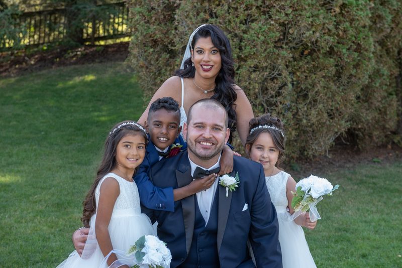A bride and groom smile with three young flower children in matching outfits as sunlight filters through trees.