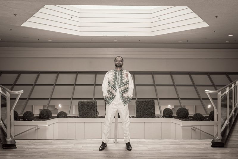 A man in white and a patterned vest stands in a modern atrium with geometric skylights and railings.