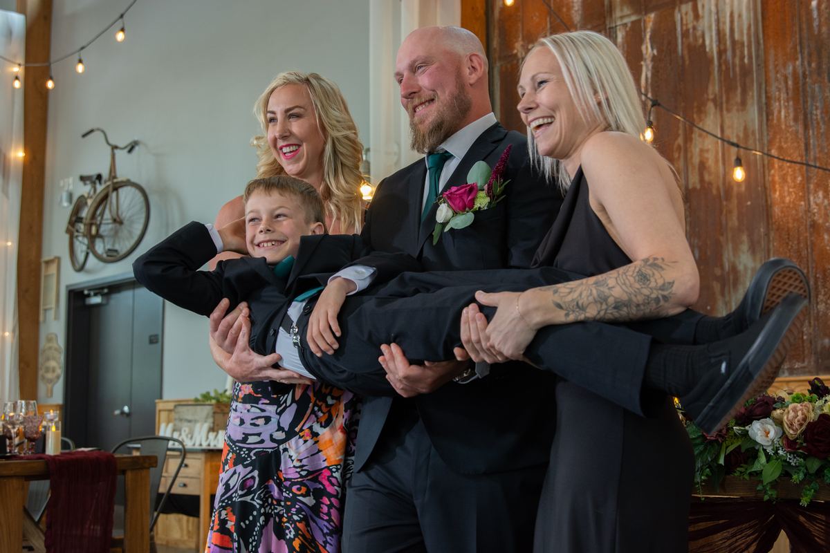 A groom and two brides smiling and laughing as they carry a joyful young boy at what seems to be a wedding reception.