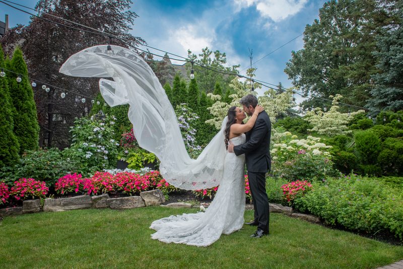 A bride and groom kiss in a garden with billowing veil, vibrant flowers, and strings of lights overhead.