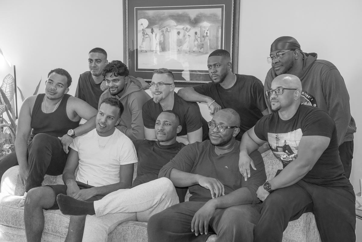 Nine men pose closely together on a sofa in a warm, intimate setting with natural light from the left.