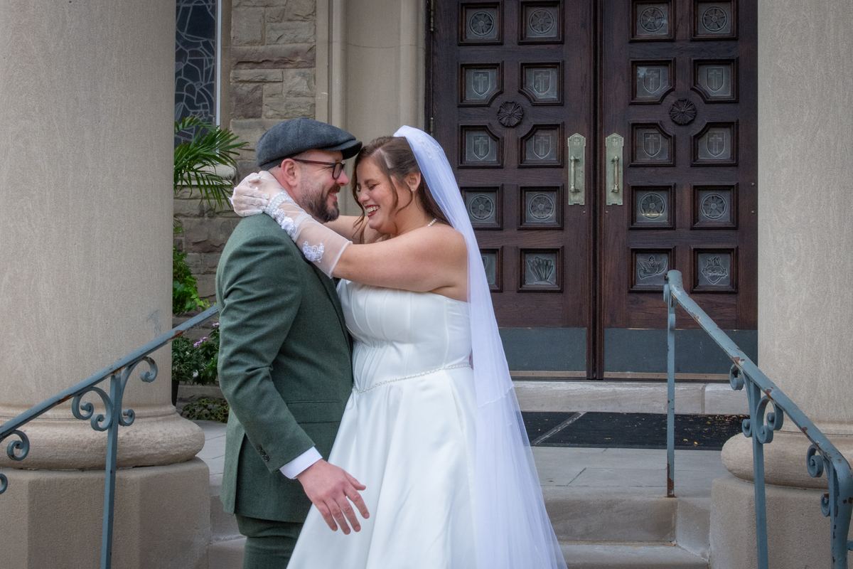 A laughing bride in a white gown and veil embraces her groom in a green suit and flat cap on stone church steps.