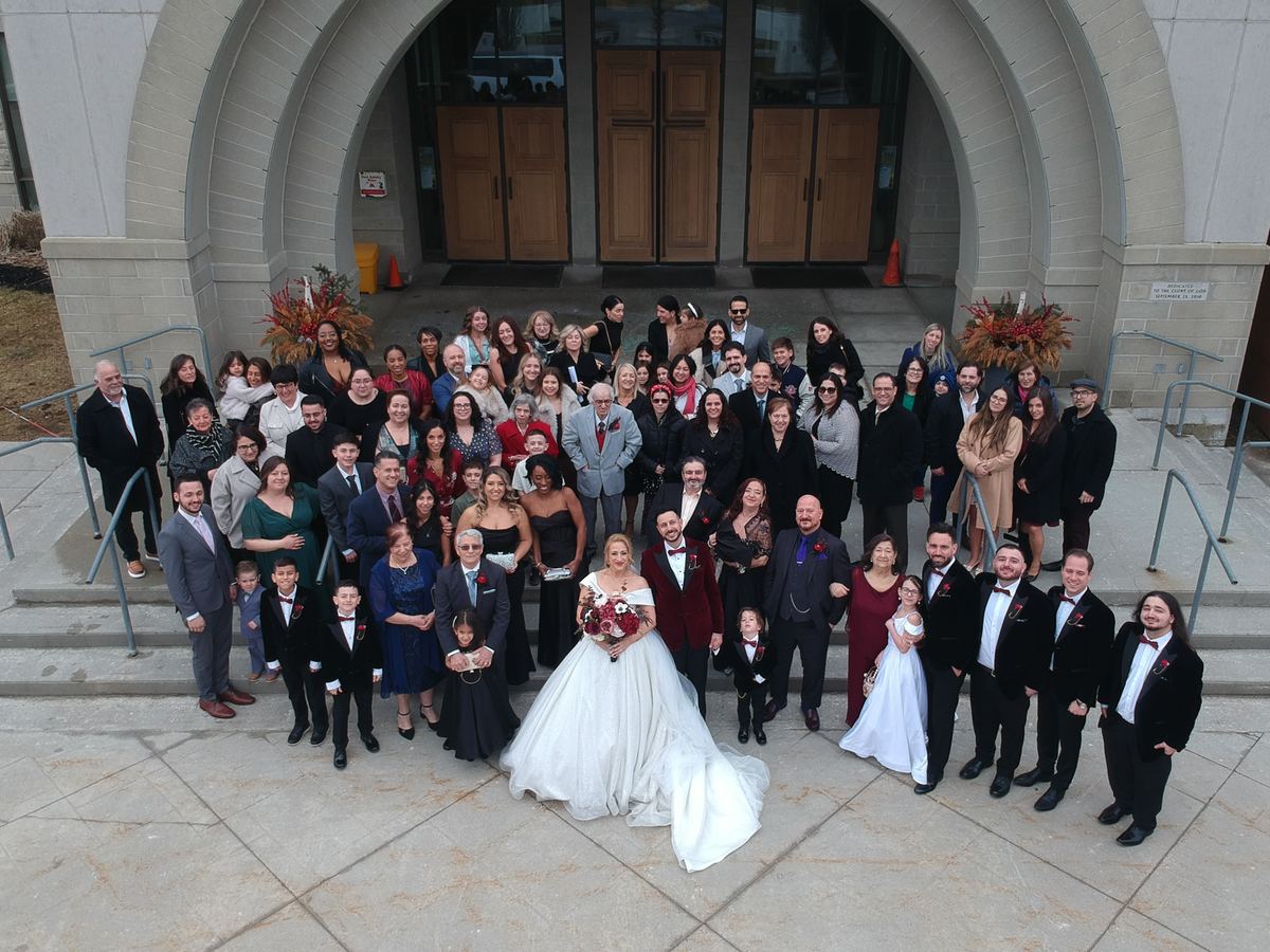 A bride and groom stand with family and friends in front of a modern stone building during an overcast day.