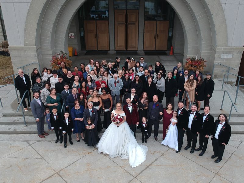 A bride and groom stand with family and friends in front of a modern stone building during an overcast day.