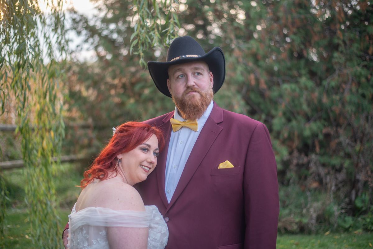 A red-haired bride in a white gown leans against her groom in a burgundy suit under weeping willow trees.