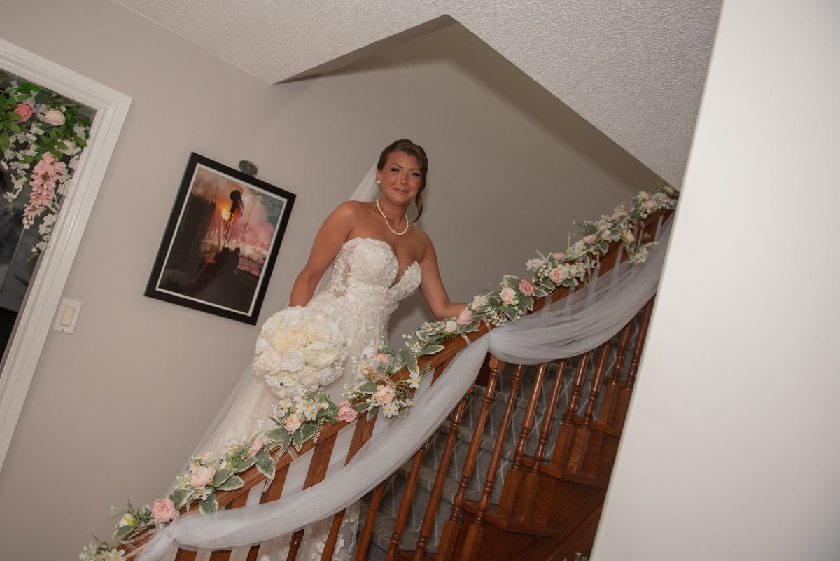 A bride in a white gown and veil stands on a wooden staircase adorned with floral garlands and fabric.