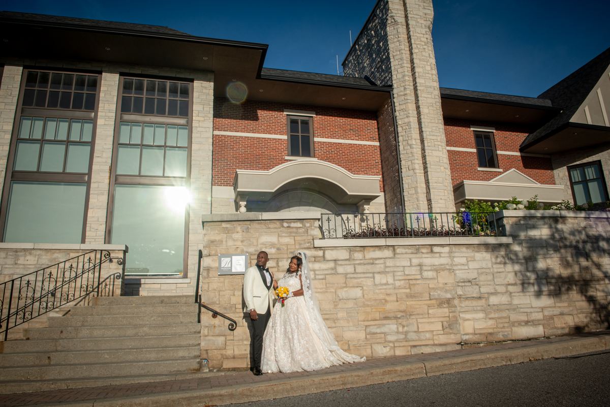 A newlywed couple in formal attire stands on stone steps under bright sunlight, the bride holding a bouquet.