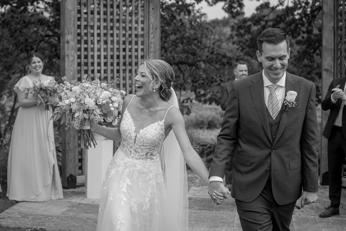 A smiling groom and laughing bride walk hand-in-hand down an outdoor aisle, guests visible in the background.