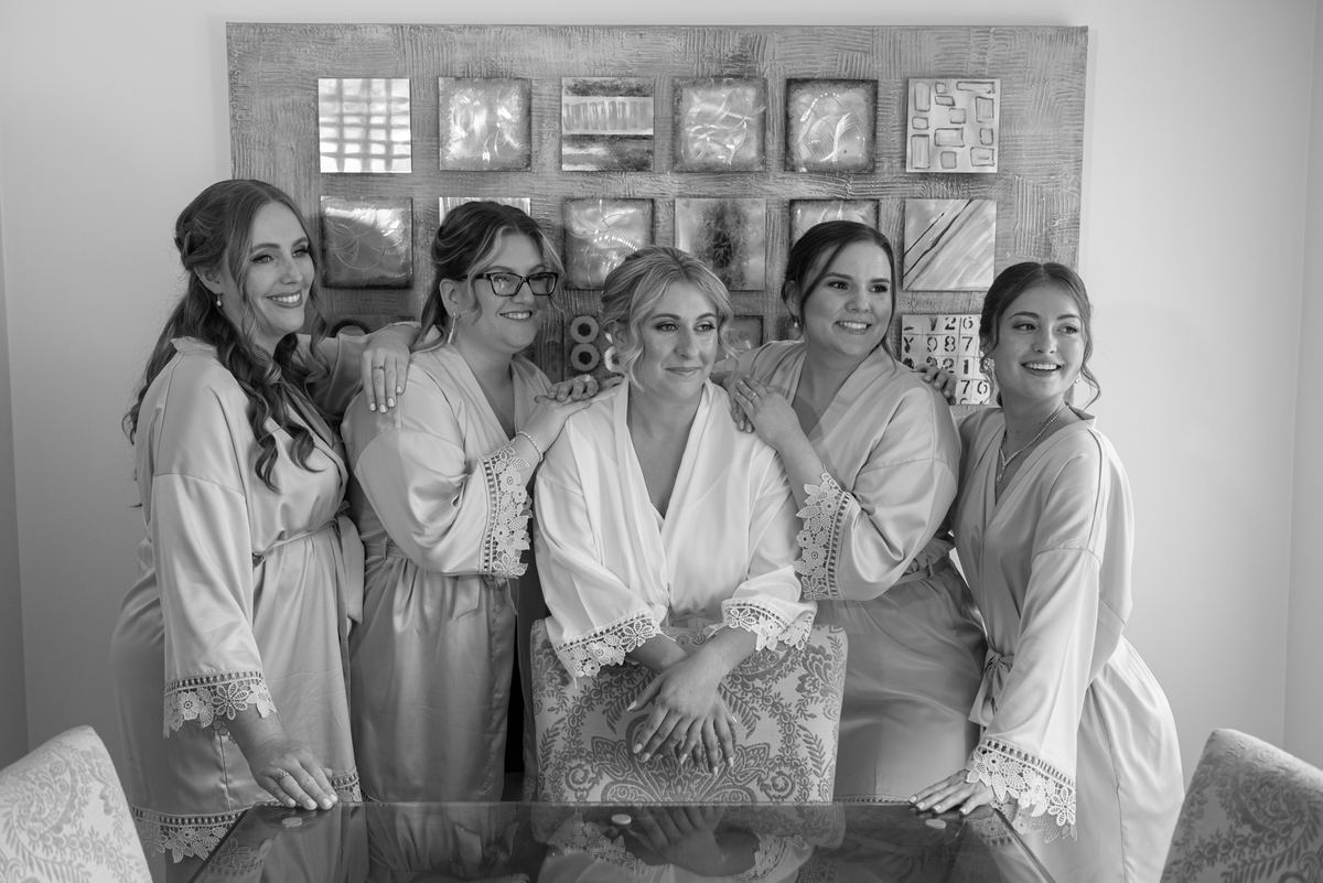 A bride and her bridesmaids smile around a glass table in matching robes during pre-wedding preparations.