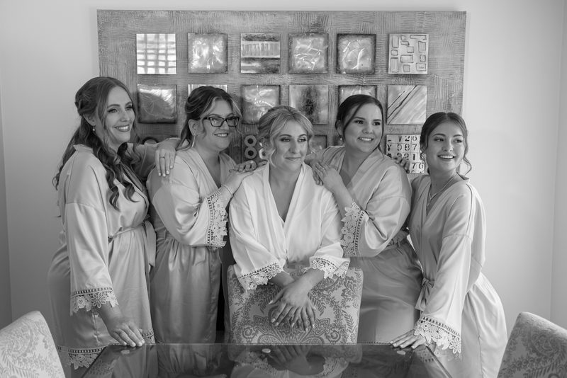 A bride and her bridesmaids smile around a glass table in matching robes during pre-wedding preparations.