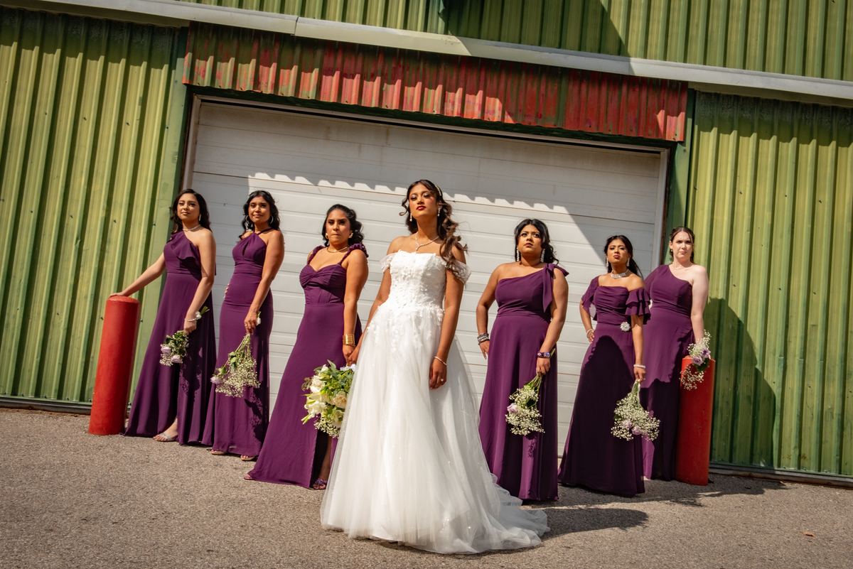 A bride and six bridesmaids stand in front of a green garage door, contrasting elegant dresses with industrial setting.