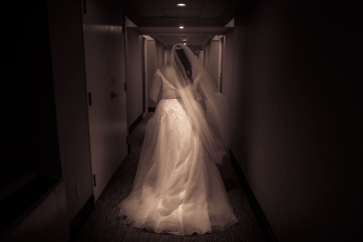 A bride in a white wedding gown and veil walks down a dimly lit corridor, her figure glowing softly under ambient lights.