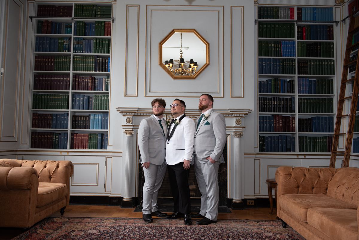 Three men in formal wear stand in a warmly lit, book-lined library with golden trim and rich wood tones.
