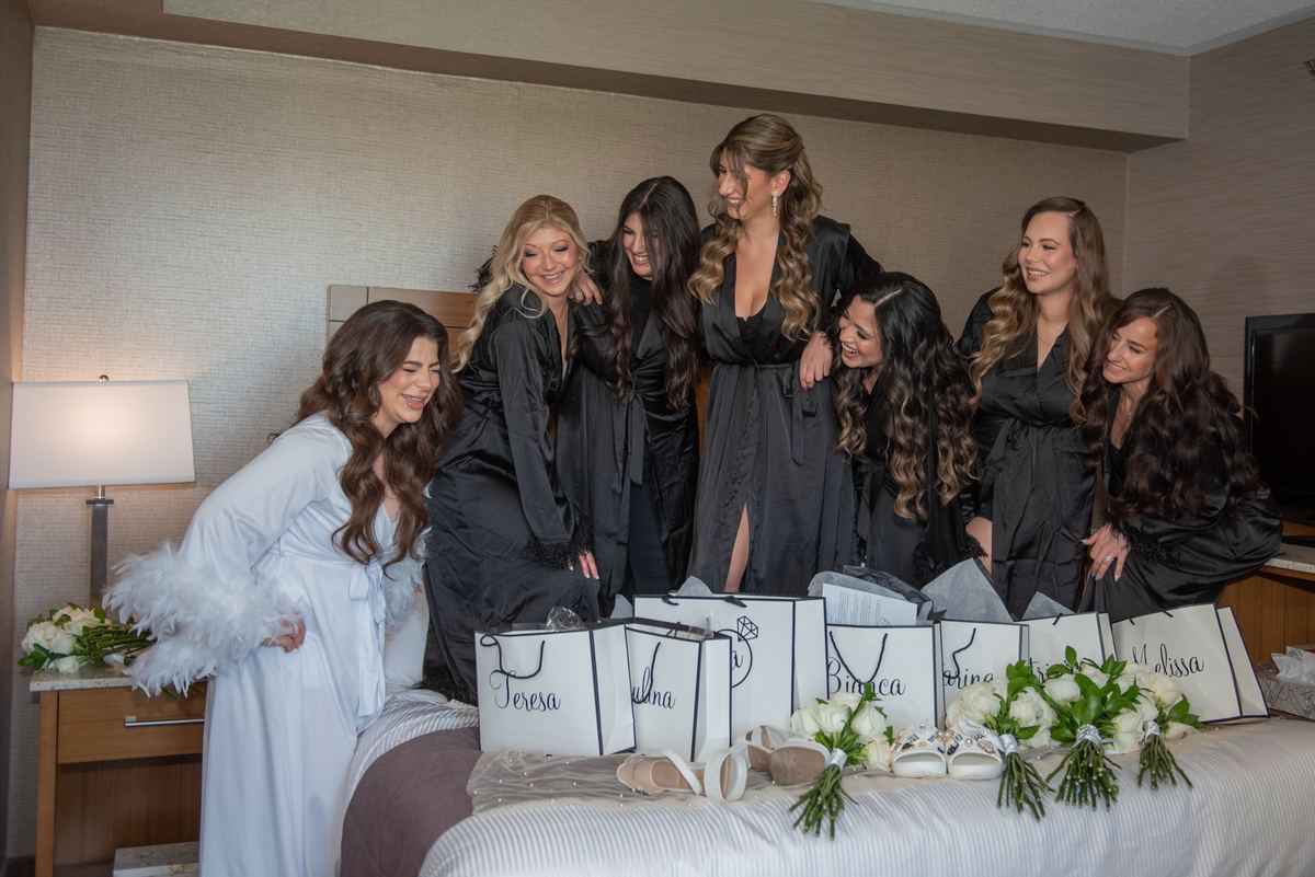 A bridal party gathers around a bed, laughing and admiring gift bags and flowers, with the bride in a white robe.