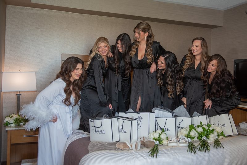 A bridal party gathers around a bed, laughing and admiring gift bags and flowers, with the bride in a white robe.