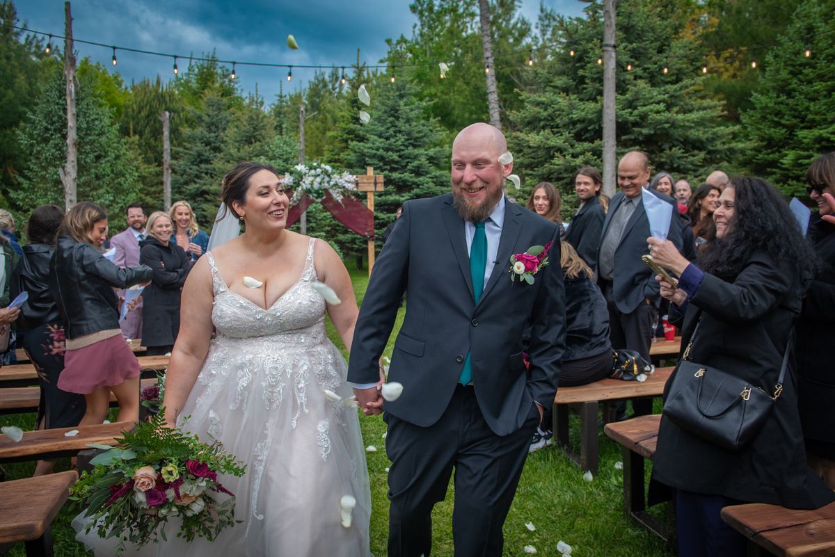 A smiling bride and groom walk down an aisle surrounded by guests tossing flower petals, under string lights.