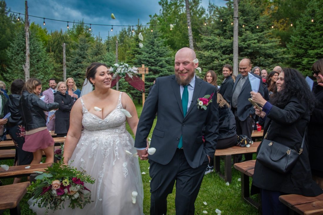 A smiling bride and groom walk down an aisle surrounded by guests tossing flower petals, under string lights.