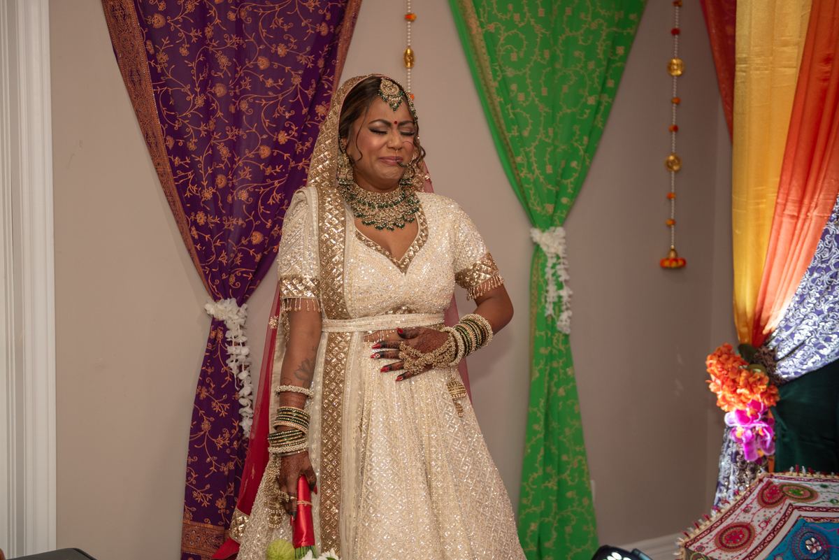 A bride in an ornate cream lehenga with gold embroidery and jewelry stands smiling at a colorful South Asian wedding.