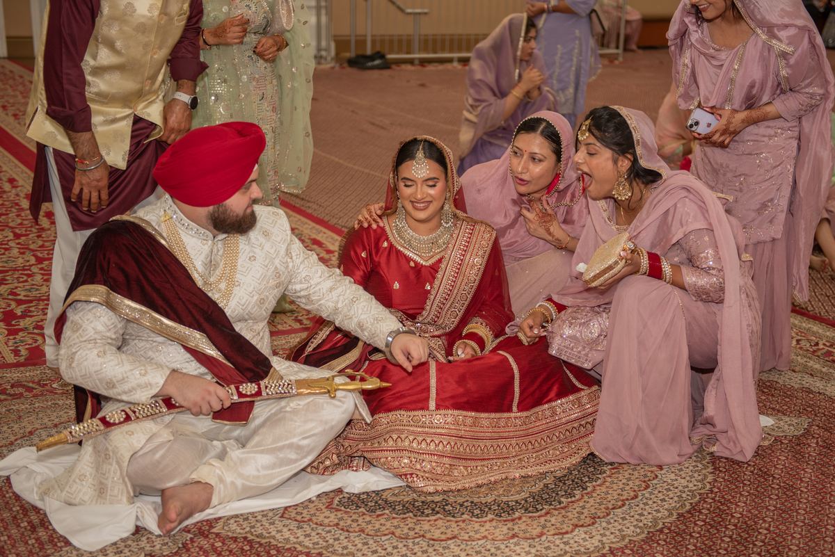 A Sikh groom and his bride sit surrounded by smiling female relatives in a decorated hall during a joyful celebration.