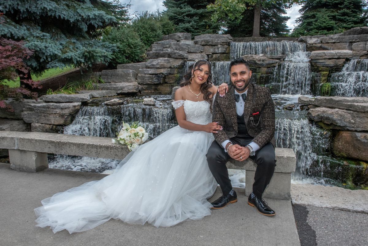 A newlywed couple poses joyfully on a stone bench beside a multi-tiered waterfall, the bride in a white gown and bouquet.