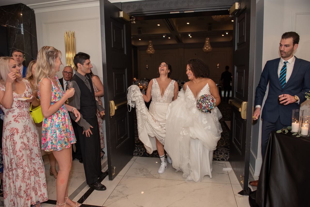 Two brides laughing as they enter a reception hall, dragging their gowns, with guests cheering from the doorway.