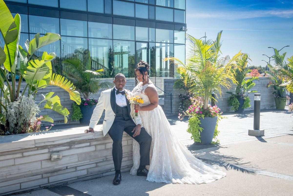 A bride in white and groom in tuxedo pose joyfully beside a modern glass building, framed by lush plants and blue sky.