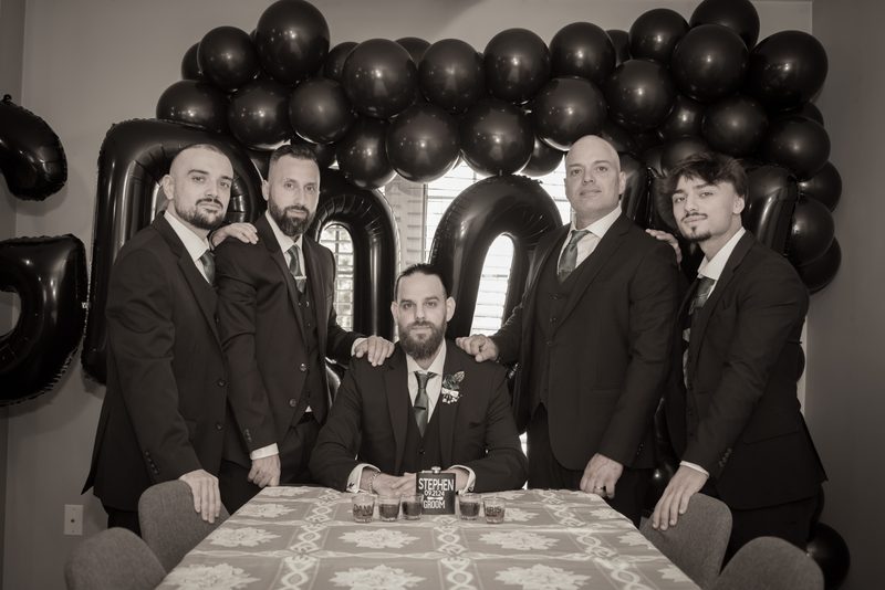 A formally dressed groom and four standing groomsmen smile at the camera with a '20' balloon arch behind them.