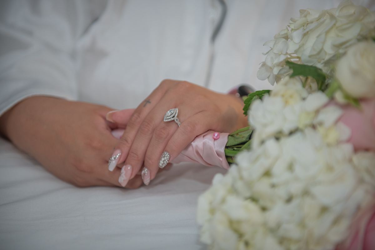 Bride and groom hold hands, one wearing a diamond ring, white hydrangeas nearby, soft light highlighting details.