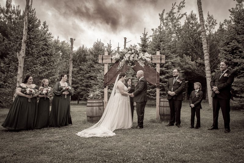 Bride in white and groom in dark suit stand under wooden arch adorned with flowers during outdoor ceremony.