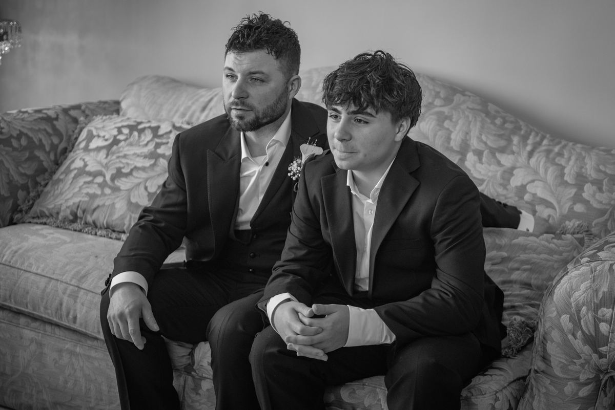 Two formally dressed men sit pensively on an ornate sofa, their expressions focused out of frame in a softly lit scene.
