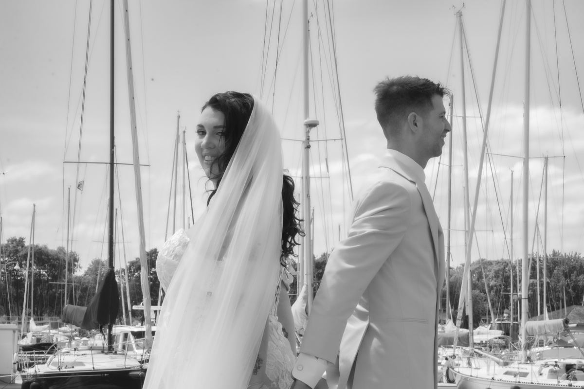 A newlywed couple walks along a marina, the bride in a veil smiling at the camera while the groom looks ahead.
