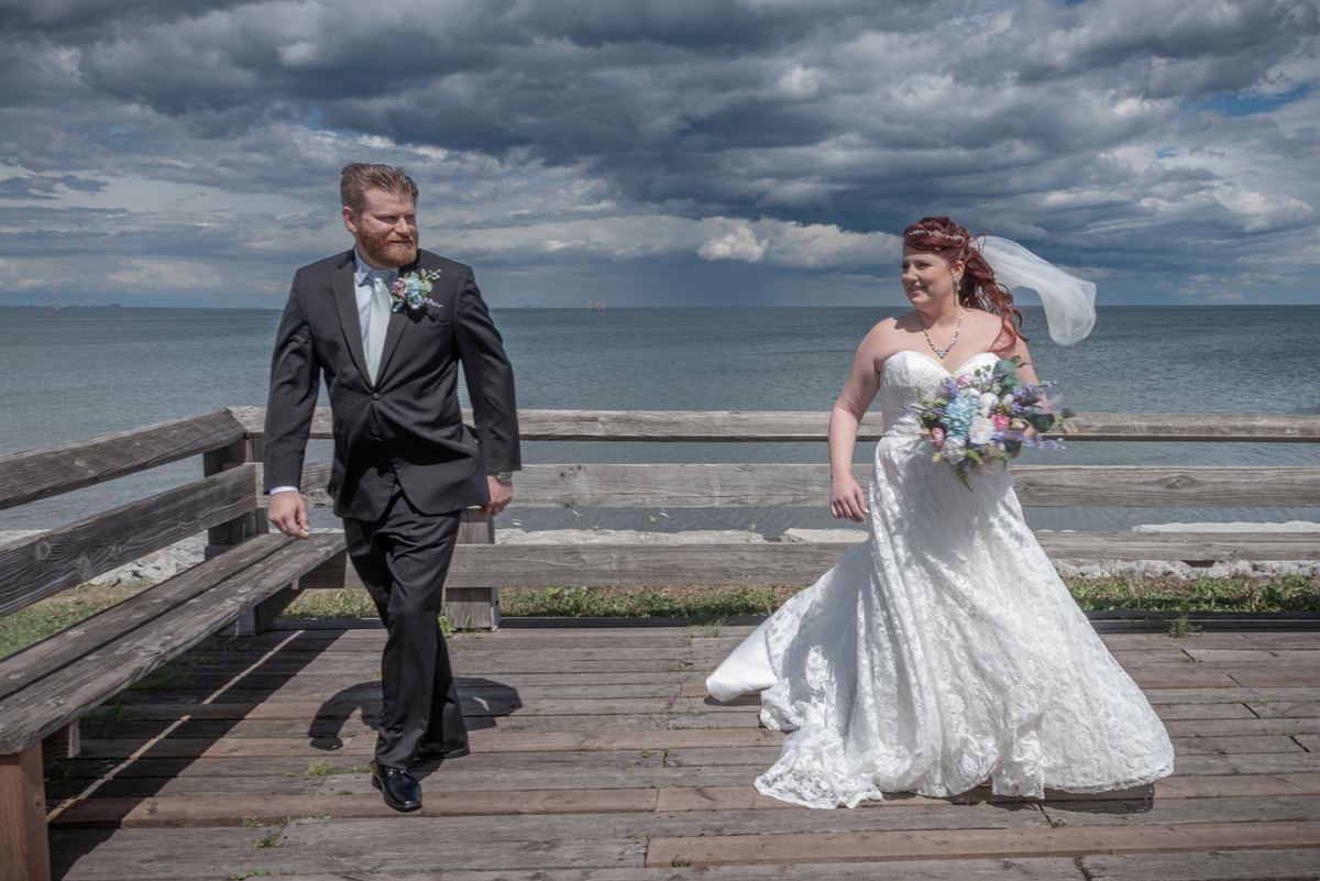 Meghan and Will stroll side by side along a wooden pier, the bride smiling as she holds her bouquet.