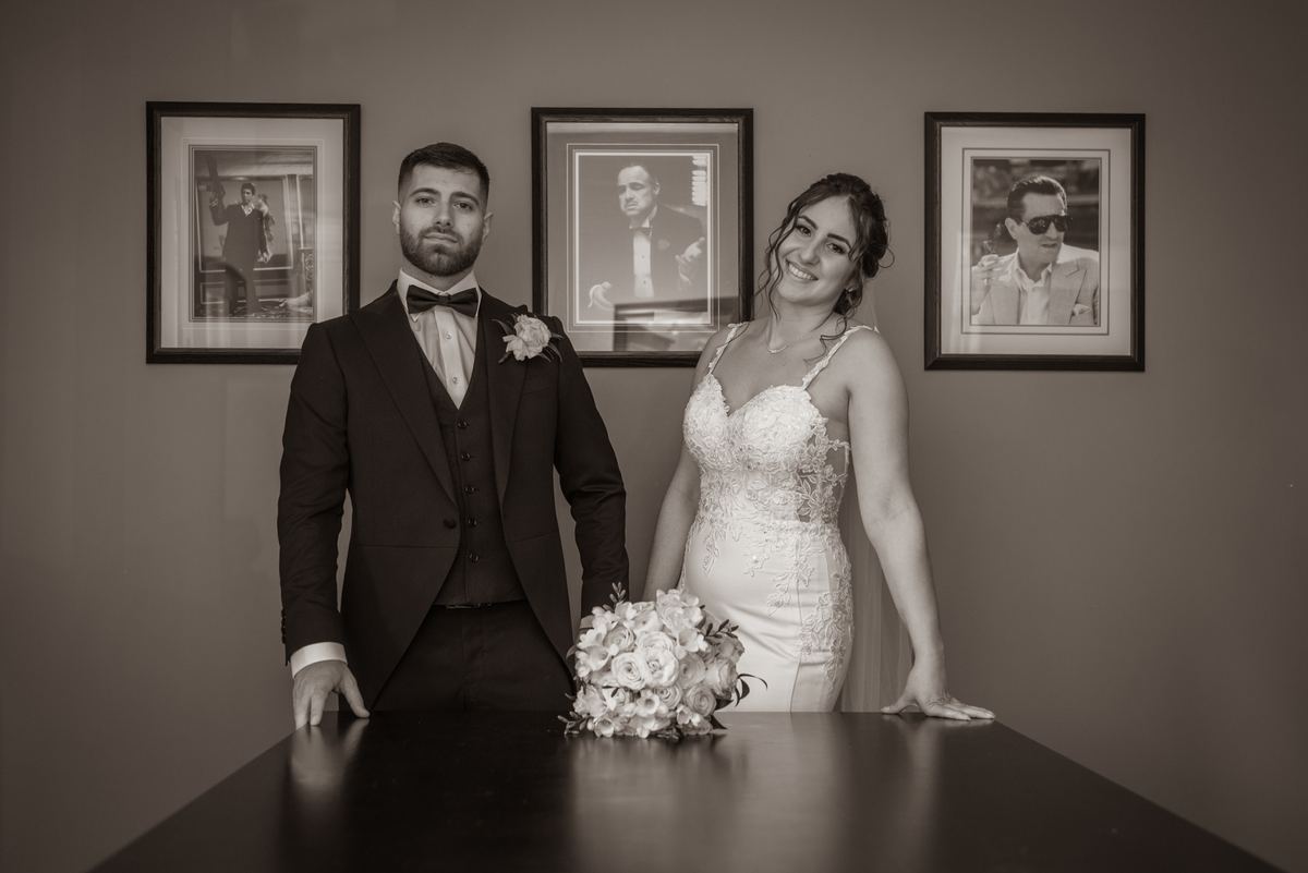 A newlywed couple poses formally at a dark table with soft lighting and framed portraits behind them.