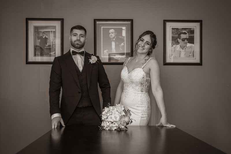 A newlywed couple poses formally at a dark table with soft lighting and framed portraits behind them.