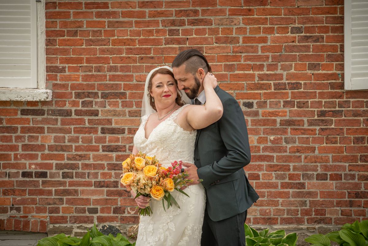 A bride and groom embrace by a red brick wall, the bride's bouquet of yellow and orange flowers visible in her hand.