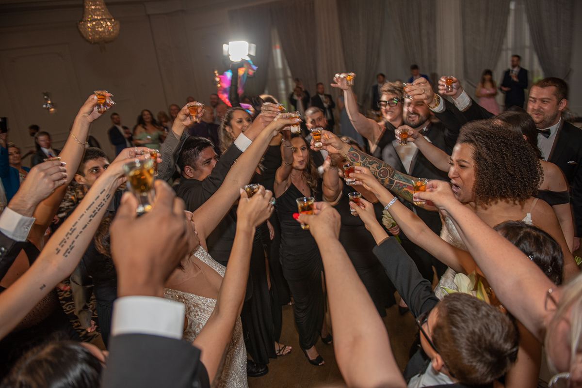 Guests in formal attire raise shot glasses and cheer at a wedding reception, illuminated by warm lights and camera flash.