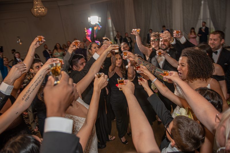 Guests in formal attire raise shot glasses and cheer at a wedding reception, illuminated by warm lights and camera flash.