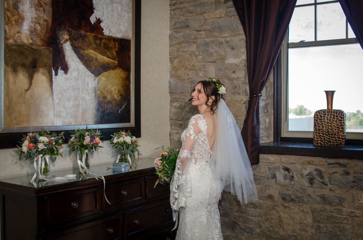 A smiling bride in an elegant lace gown and veil stands by a stone wall, gazing at the light filtering through a window.