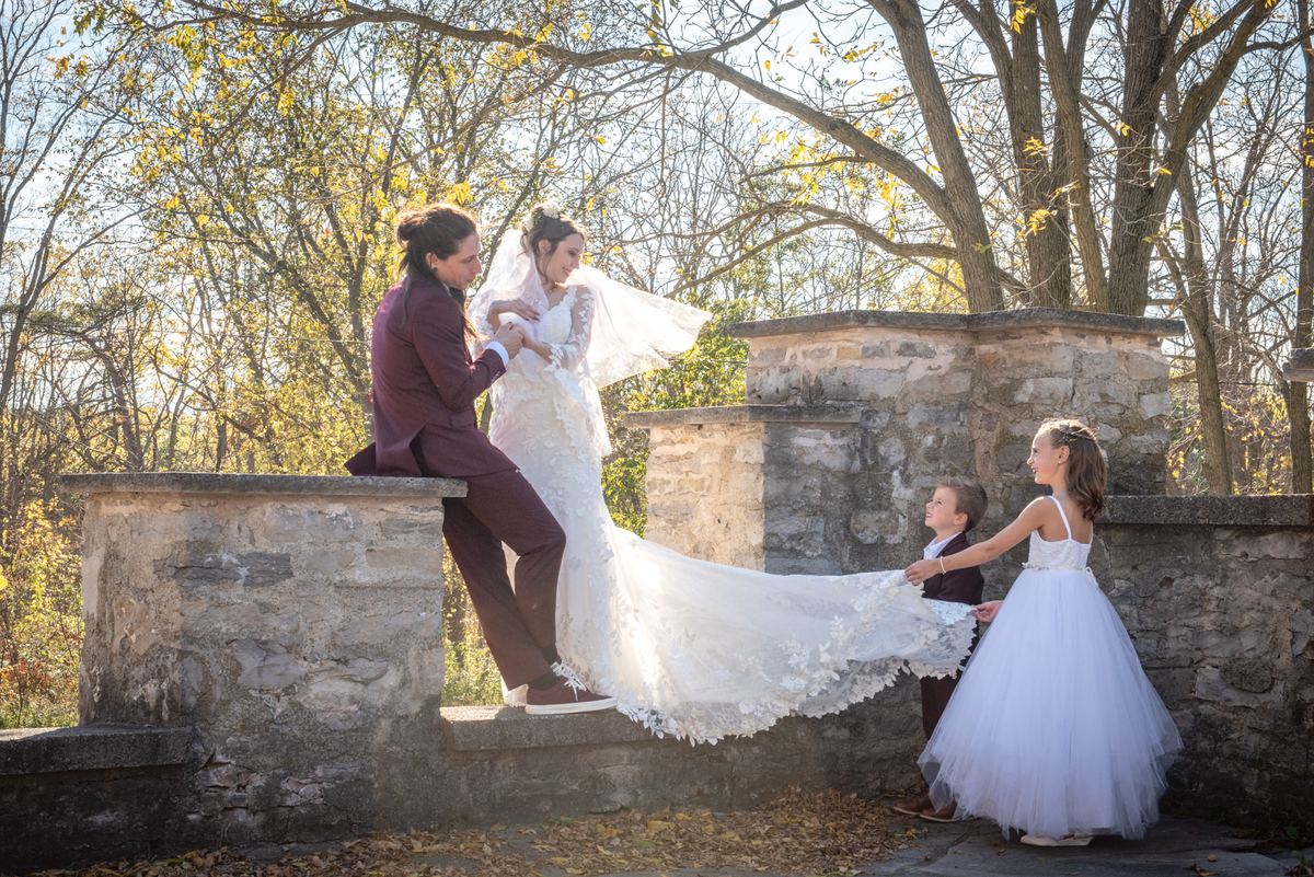 Bride in white gown steps down from stone wall with groom and children holding train, sunlight filters through trees.