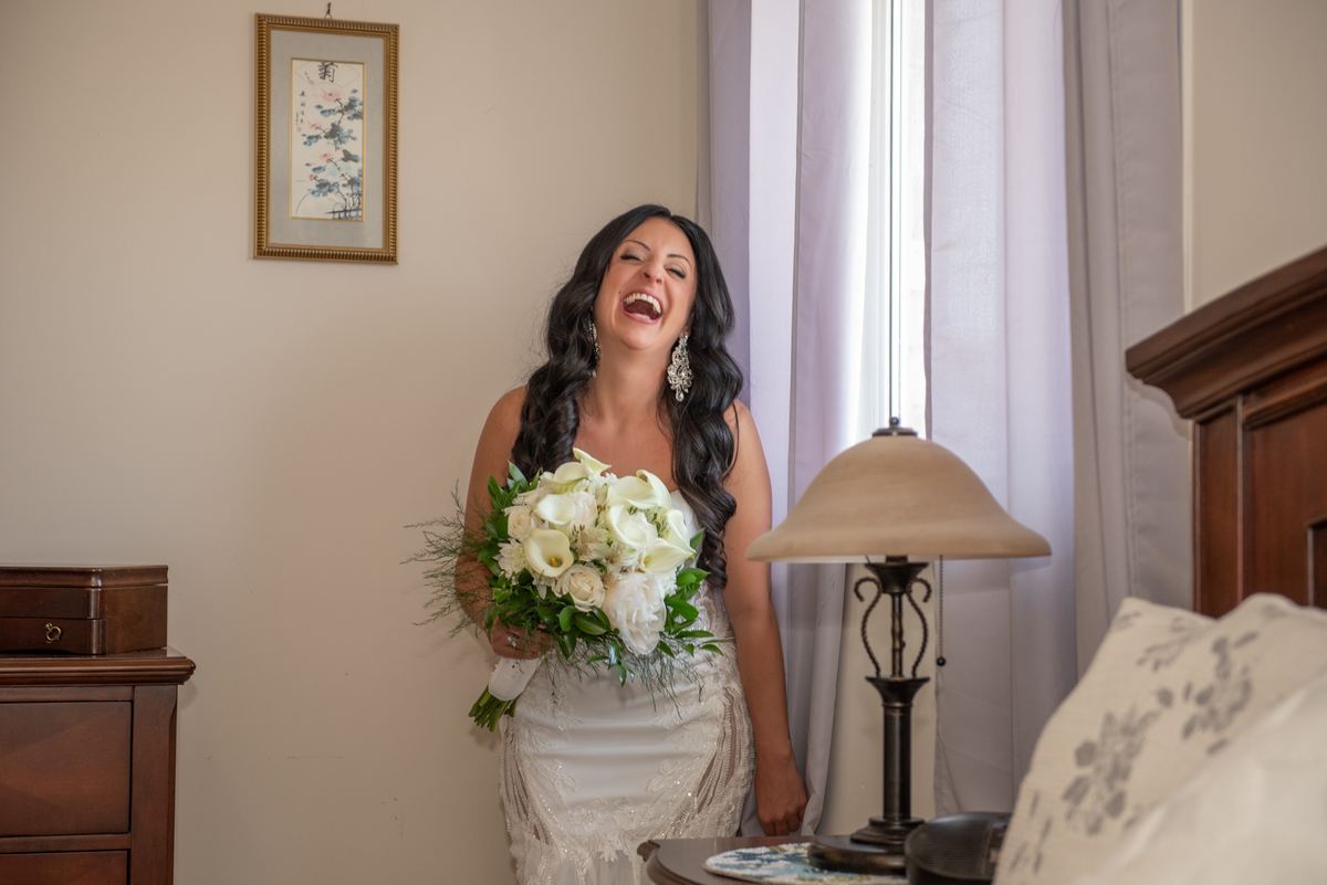 A bride in a white gown holding flowers laughs near a window with sheer curtains and a wooden dresser.